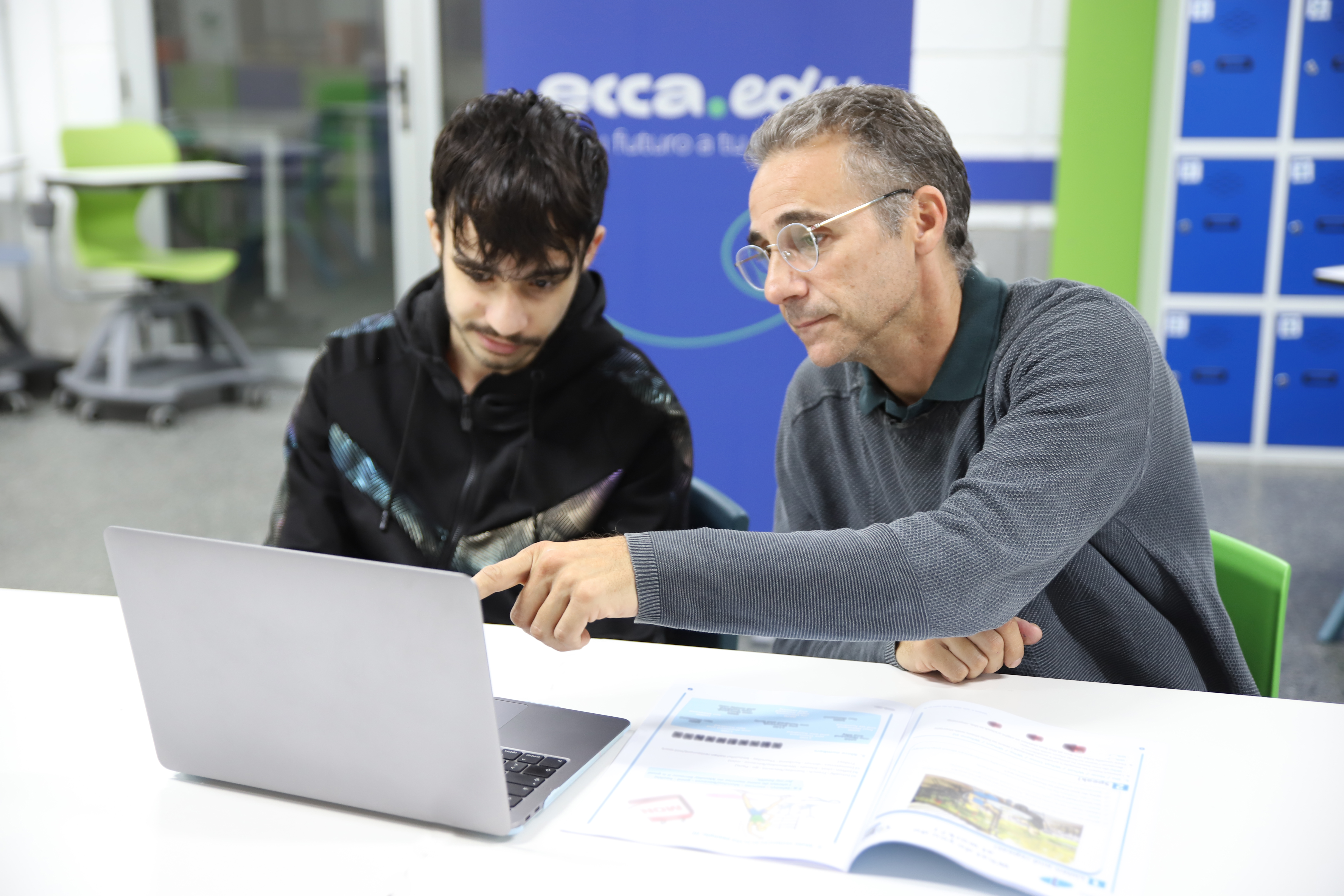 Un profesor sentado junto a un alumno señala la pantalla de un portátil con un módulo de ecca.edu abierto en la mesa